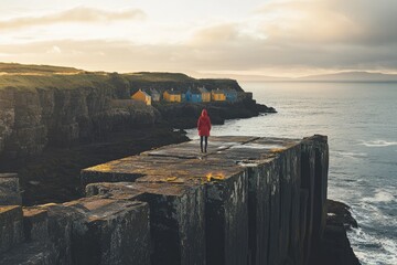 Solitary figure in red coat stands on a cliff overlooking colorful houses by the sea at sunset.
