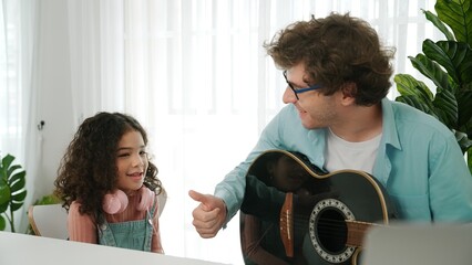Skilled caucasian father playing guitar to relax daughter after learning engineering prompt and code. Cute schoolgirl with headphone sitting at table with laptop while listen acoustic music. Pedagogy.