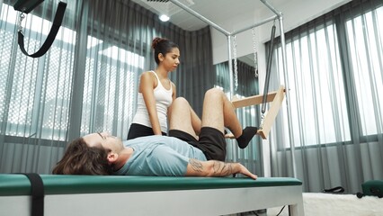 Skilled pilates instructor assisting a client in leg lift exercise on Cadillac equipment. Female trainer assist leg lift stretch on cadillac for improving core engagement, leg flexibility. Habituate.