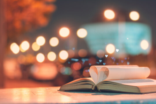 Open book with pages folded into a heart shape, placed on a table with a dreamy bokeh background of warm city lights at night, evoking a romantic and literary mood.
