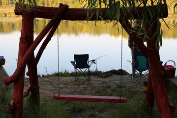 A tranquil lakeside scene with a red swing, chairs, and fishing gear.