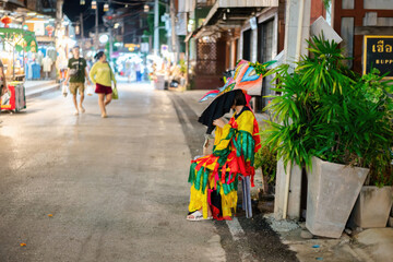 Fototapeta premium Vibrant street scene featu a woman in colorful traditional dress sitting under a black umbrella next to lush green potted plants on a lively city sidewalk at night