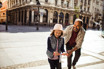 Father teaching daughter to ride scooter on sunny city street