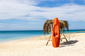 Orange colored canoe rests in a standing position on a white sand beach with a calm blue sea...