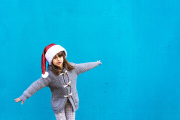 joyful girl in santa hat spreading arms in front of a bright blue wall. Her playful expression and festive outfit capture the spirit of childhood fun. Christmas holiday and generation alpha concept.