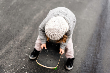 Defocused portrait of little girl in winter wool clothes while playing with skateboard on the road against asphalt background - Childhood and sport concept - Main focus on the wool hat