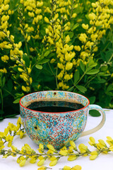A floral ceramic cup with freshly brewed black coffee sits on a garden table with bright yellow...
