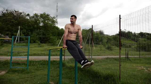 Strong man trains abdominal muscles raising legs on parallel bars at sports ground. Young shirtless sportsman exercising at nature. Sporty athlete working out at nature. Concept of sportive lifestyle