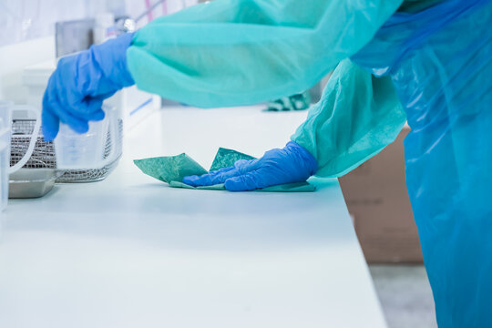 A close-up view of a healthcare professional in protective medical scrubs and blue sterile gloves meticulously cleaning a pristine white laboratory counter with a green cloth, prioritizing hygiene