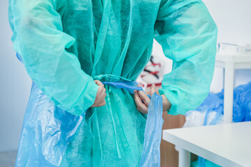 View from behind as a healthcare professional ties a blue disposable plastic apron over their green medical gown, emphasizing infection control