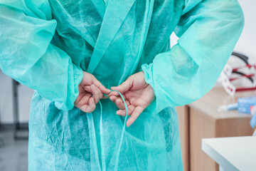 Close-up back view of a medical worker's hands tying the light green disposable isolation gown,...