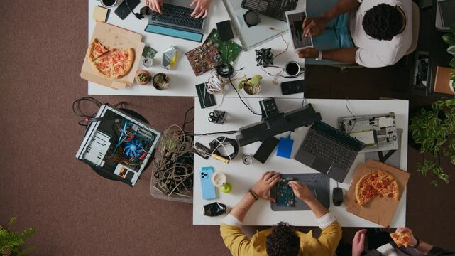 Directly above shot of technician fixing circuit board when colleague eating pizza and helping him with advice standing near table, programmer using digital tablet, unknown person typing on laptop