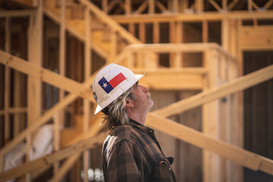 Construction Worker Wearing Hard Hat with Texas Flag Sticker Walking Inside of and Looking Up at Wooden Frame of House at Construction Site