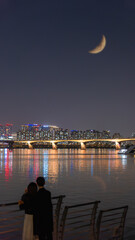 A couple gazes at the illuminated bridge and city lights from Yeouido Park, Seoul, under a crescent...