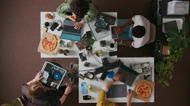 Directly above shot of four diverse programmers eating pizza, using tablet and laptops, taking cables out of container during workday at white desks in office