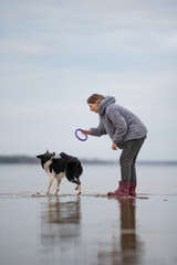 A woman wearing a grey jacket and red boots is playing with her black and white Border Collie dog on a beach next to the water. She is holding a ring toy and about to throw it