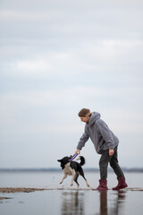 A woman in boots and a grey hoodie plays tug of war with a black and white Border Collie using a...