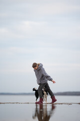 A young woman is carefully walking and balancing on a thin strip of beach with water on either side. A black and white border collie follows behind her on a cloudy day