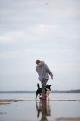 A young woman is standing in shallow water near the sea, with a border collie dog standing on her foot inside her Wellington boot. The sky is overcast