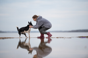 A woman in a grey jacket and red boots kneels at the water's edge, gently holding a black and white Border Collie. The water is still, creating a mirror-like reflection of the pair