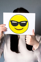 Woman holding happy emoji with sunglasses mask in front of face. Making thumbs up. World emoji day...