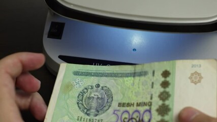 Examination of Uzbekistan Sum (UZS) cash money with UV counterfeit detector. Concept of authenticity check, fight against counterfeiting of banknotes, anti fraud measures in Uzbekistan. Close up view.