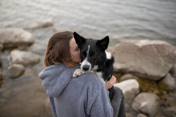 A woman in a grey hooded top is hugging a black and white Border Collie dog. They are standing on a...