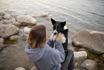 A woman in a hooded sweatshirt sits on rocks at the water's edge. She is holding a border collie...