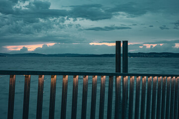 A calm evening view of Lake Constance with glowing lights shining through a metal fence. Mountains...