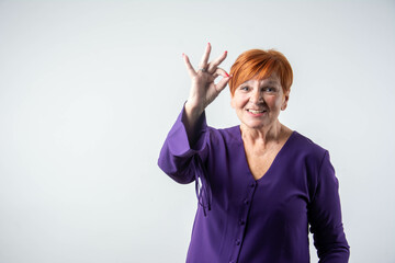 Smiling senior woman with red hair showing OK hand sign and feeling cheerful, positive emotion,...