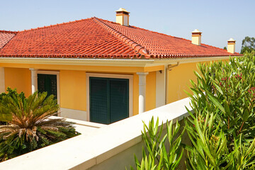 Bright Mediterranean-style house with red tiled roof, yellow walls, green shutters, and lush garden...