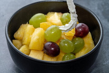 Topping fresh fruit salad with whip cream. colorful pieces of grape, orange in black bow on grey stone background, close up