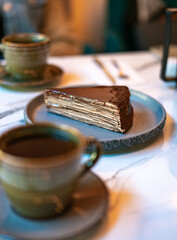 Chocolate Mille Crepe Cake Slice with Coffee Cups on Marble Table in Cozy Cafe