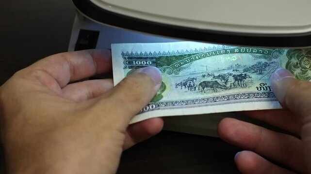 Examination of Lao Kip (LAK) cash money with UV counterfeit detector. Concept of authenticity check, fight against counterfeiting of banknotes, anti fraud measures in Laos. Close up view.
