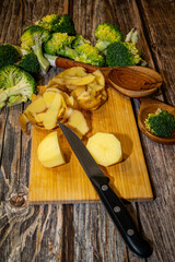A sharp knife lays on a wooden cutting board with peeled and cut potatoes with broccoli florets on...