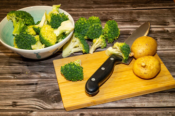 Fresh broccoli florets in a bowl and prepared on a cutting board with potatoes, ready for cooking,...