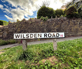 A road sign displaying "Wilsden Road 16" is set against a backdrop of a stone wall and lush greenery. The sky is partly cloudy, creating a picturesque scene on, Wilsden Road, Wilsden, Bingley, UK