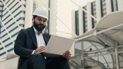 Portrait of happy Indian businessman in helmet attending online meeting via laptop in downtown. Experienced engineer talks to colleagues on video chat against commercial buildings