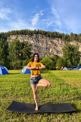 Young woman practices tree pose on a yoga mat in a summer campsite surrounded by blue tents, forest, mountains and open sky, a girl in a tree pose does yoga in nature.