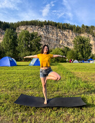 Young woman practices tree pose on a yoga mat in a summer campsite surrounded by blue tents, forest, mountains and open sky, a girl in a tree pose does yoga in nature.