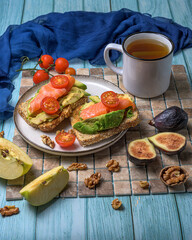 Healthy breakfast on rustic wood featuring avocado toast with salmon and cherry tomatoes, figs, walnuts, apple slices, and herbal tea in soft natural light