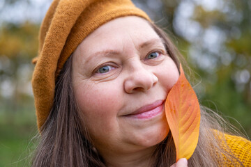 Happy mature woman in beret enjoying autumn nature, with fall leaf in hand, graceful aging, vibrant...