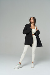 Woman wearing a cream athletic set, black jacket over shoulders, looking aside. She stands...