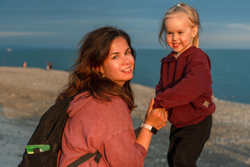 Child girl toddler with her young mother sitting on the embankment on a summer evening, people in...