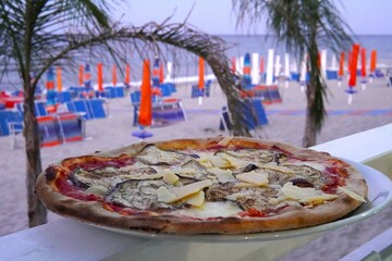 Close-up of pizza on a plate with a view of the sea and palm trees. Italian cuisine with the beauty of a tropical seaside holiday. Outdoor dining.