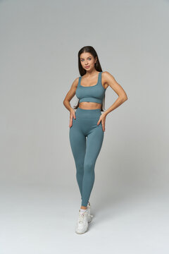 Fitness model in teal activewear, standing with a confident pose. She wears a sports bra and leggings, paired with white sneakers. Her long dark hair is sleek and straight, hands on hips