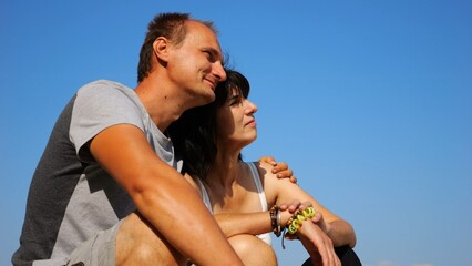 Happy pair hugs during sitting on wooden log on beach. Romantic couple admiring sea landscape at sunny day. Lovers spending time together at resort. Concept of summer vacation or holiday. Slow mo