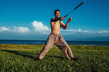 Young strong man martial arts training with nunchaku on lake shore on summer day