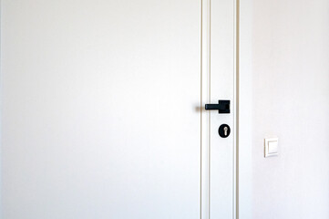 Closed white door with black handle and keyhole, next to a white light switch on a plain wall