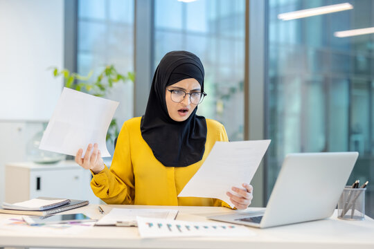 A shocked businesswoman in a hijab reviews paperwork in an office setting. The image portrays surprise and potential work-related stress.